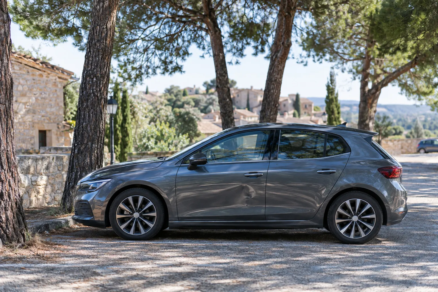Voiture garée sur un petit parking de village provençal bordé de pins, portière légèrement cabossée